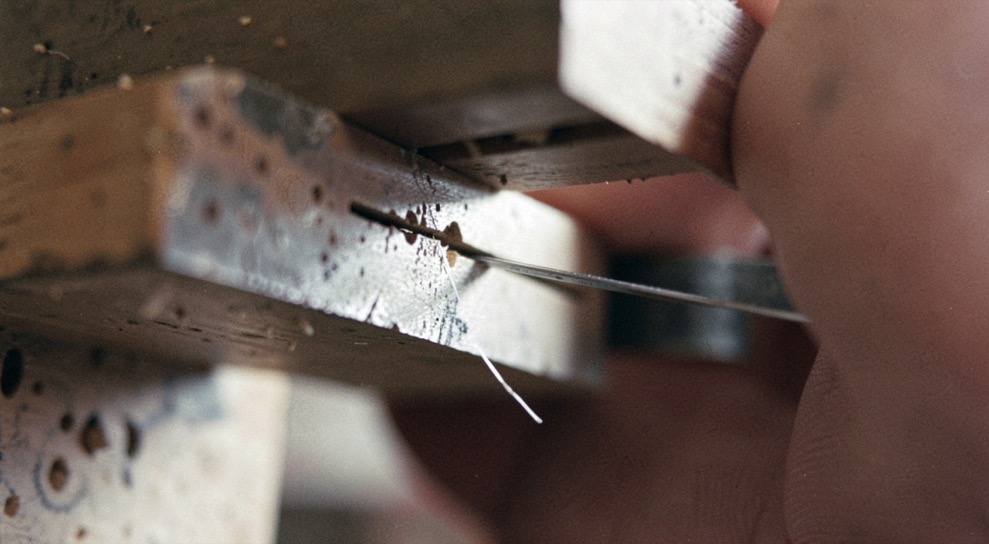 A very close view of the underside of a block plane showing the narrow slot between the sliding mouth plate and the blade edge, a thin shaving emerging, hard task light from one side.